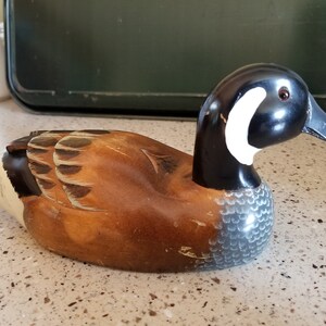 May include: A wooden carved duck decoy with brown, black, and white paint. The duck has a black head with a white patch on the side and a black beak. The body is brown with a gray and white pattern on the bottom.