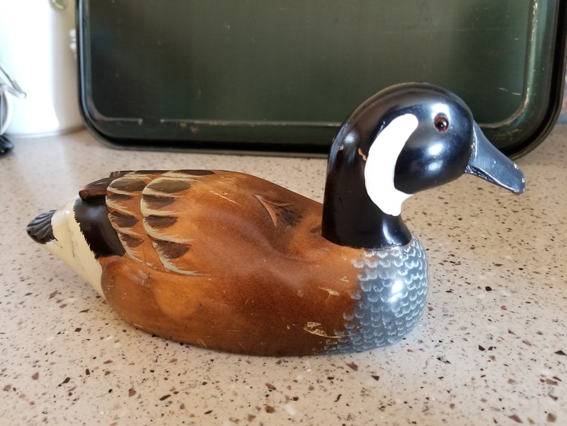 May include: A wooden carved duck decoy with brown, black, and white paint. The duck has a black head with a white patch on the side and a black beak. The body is brown with a gray and white pattern on the bottom.