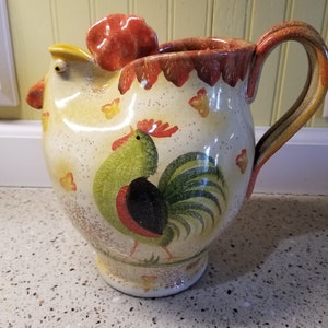 May include: A ceramic pitcher shaped like a rooster with a white background and a rooster painted in green, red, and black. The pitcher has a handle and a spout.