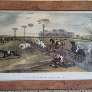 May include: A vintage print depicting a steeplechase race in the Vale of Aylesbury. The print shows several horses and riders jumping over a fence, with one horse and rider having fallen. The print is framed in a wooden frame.