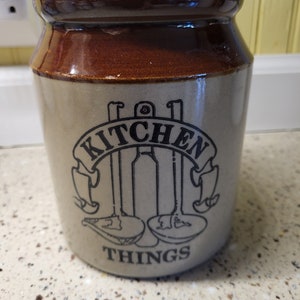 May include: A brown and beige ceramic kitchen utensil holder with the words "Kitchen Things" and a graphic of kitchen utensils.