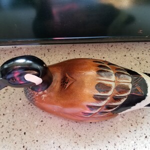 May include: A wooden carved duck decoy with brown, black, and white paint. The duck has a black head with a white stripe on the side. The body is brown with black and white markings.