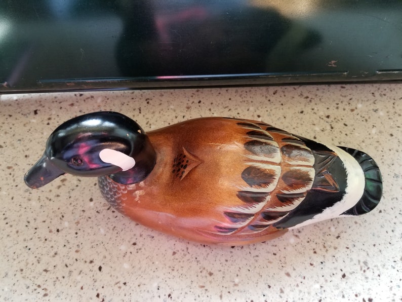 May include: A wooden carved duck decoy with brown, black, and white paint. The duck has a black head with a white stripe on the side. The body is brown with black and white markings.