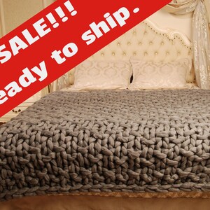 May include: A gray chunky knit throw blanket on a bed with white bedding and a tufted headboard. The blanket is made of thick yarn and has a textured weave.