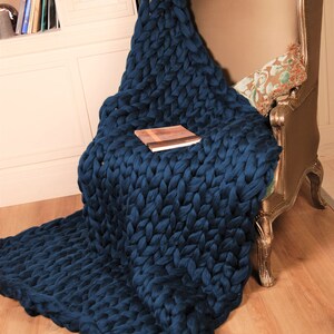 May include: A large, chunky knit throw blanket in a deep blue color draped over a chair. The blanket is made of thick yarn and has a soft, cozy texture.