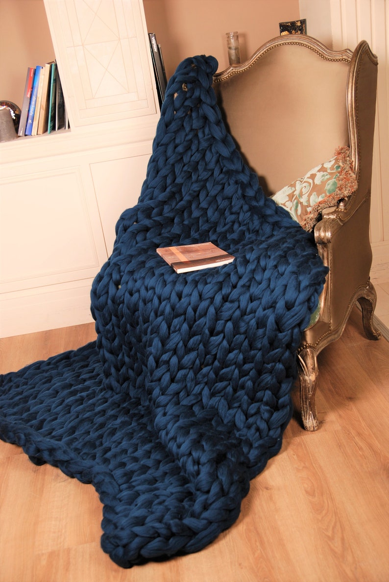 May include: A large, chunky knit throw blanket in a deep blue color draped over a chair. The blanket is made of thick yarn and has a soft, cozy texture.