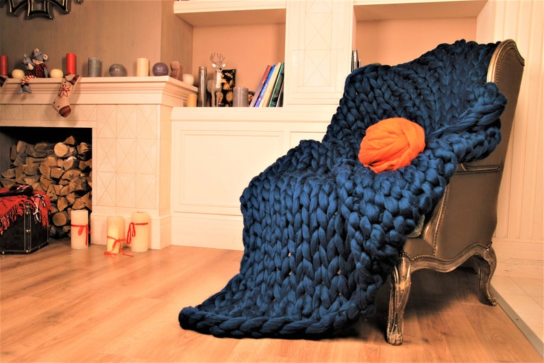 May include: A large, chunky knit throw blanket in a deep blue color draped over a chair. The blanket is made of thick yarn and has a soft, cozy texture.