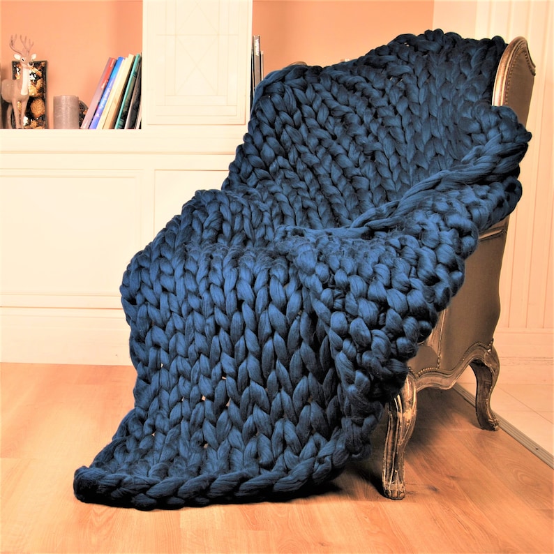May include: A large, chunky knit throw blanket in a deep blue color. The blanket is draped over a chair, showcasing its soft and cozy texture.