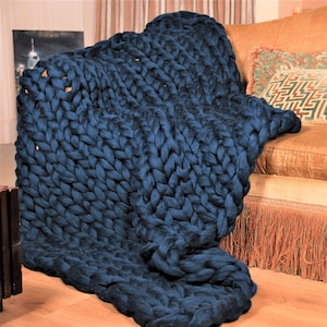 May include: A large, chunky knit throw blanket in a deep blue color. The blanket is draped over a couch with a fringe trim.