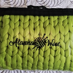 May include: A bright green chunky knit blanket in a clear plastic bag with a black drawstring bag. The blanket has a logo that says "Alanna Wool" with a stylized yarn ball and needle.