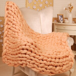 May include: A chunky knit throw blanket in a peach color draped over a white armchair. The blanket is made of thick yarn and has a soft, cozy texture.