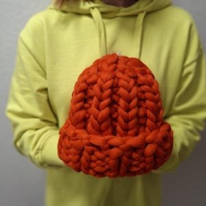 Peut inclure: Un bonnet en tricot épais de couleur orange vif. Le bonnet est fait de laine épaisse et a une coupe ample.