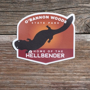 May include: A sticker featuring the text "O'BANNON WOODS STATE PARK" and "HOME OF THE HELLBENDER". The sticker has a brown and orange background with a black silhouette of a salamander.