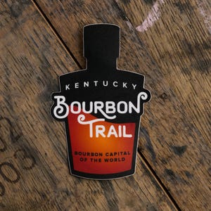 May include: A black sticker in the shape of a bourbon bottle. The sticker reads "Kentucky Bourbon Trail" in white and red text. The bottom of the sticker says "Bourbon Capital of the World". The sticker is on a wooden surface.