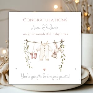 May include: A square greeting card with the words "CONGRATULATIONS" and "Anna and Jamie" in a script font. The card features a watercolor illustration of baby clothes hanging on a clothesline, with the text "You're going to be amazing parents!"