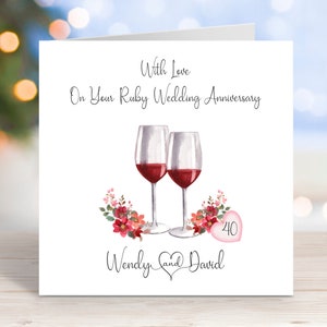 May include: A white card with a red heart and the number 40 on it. The card says "With love on your Ruby Wedding Anniversary" and "Wendy and David". There are two wine glasses with red wine in them and pink flowers.