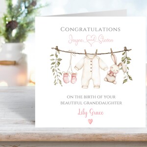 May include: A white card with the words "Congratulations Jayne and Steven" in pink script. The card features a watercolor illustration of baby clothes and a stuffed bunny hanging on a clothesline. The text "On the birth of your beautiful granddaughter Lily Grace" is also included.