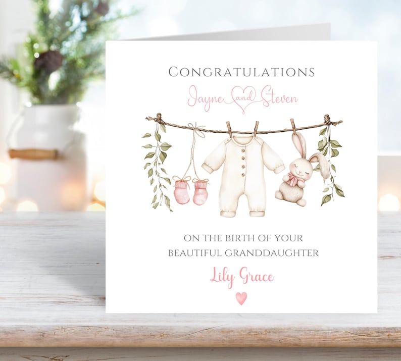 May include: A white card with the words "Congratulations Jayne and Steven" in pink script. The card features a watercolor illustration of baby clothes and a stuffed bunny hanging on a clothesline. The text "On the birth of your beautiful granddaughter Lily Grace" is also included.