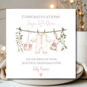 May include: A square greeting card with the words "Congratulations" and "On the birth of your beautiful granddaughter". The card features a watercolour illustration of baby clothes, a bunny, and the names "Jayne and Steven" and "Lily Grace".
