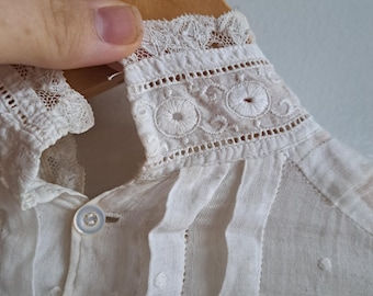 May include: White cotton blouse with a lace collar and embroidered details. The collar has a scalloped edge and the blouse has a button-up front.