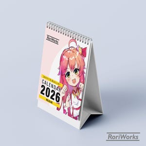 May include: A white desk calendar with a spiral binding. The calendar features a pink background with an anime character illustration. The text "CALENDAR 2026" is displayed in a yellow box. The brand name "RoriWorks" is visible.