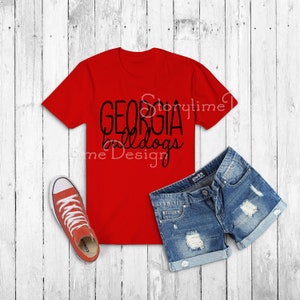May include: Red t-shirt with black text that reads "GEORGIA bulldogs". The shirt is on a white wood background with a pair of blue denim shorts and red and white sneakers.