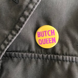 Könnte beinhalten: Ein runder, gelber Button mit dem Text "BUTCH QUEEN" in leuchtend pinkfarbenen Buchstaben. Der Button liegt auf einem dunkelgrauen Stoff mit sichtbaren Nähten.