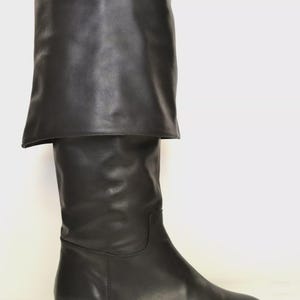 May include: A single black leather knee-high boot with a folded-over cuff. The boot has a smooth, polished finish and a low, rectangular heel. The cuff is folded down, creating a layered effect. The boot is set against a white background.