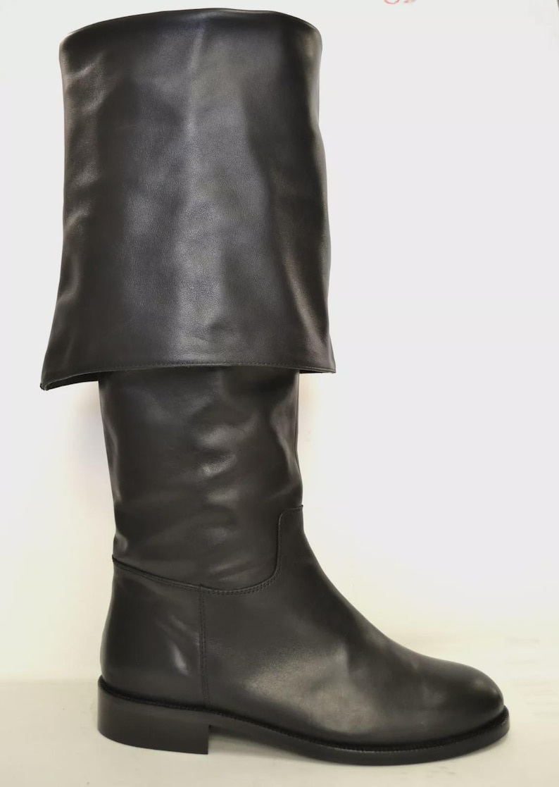 May include: A single black leather knee-high boot with a folded-over cuff. The boot has a smooth, polished finish and a low, rectangular heel. The cuff is folded down, creating a layered effect. The boot is set against a white background.