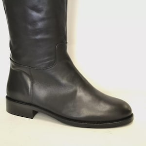 May include: A black leather knee-high boot with a low heel. The boot has a smooth, polished finish and a simple, classic design. The shaft of the boot is straight, and the toe is rounded. The heel measures approximately 1 inch.