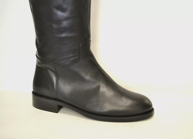 May include: A black leather knee-high boot with a low heel. The boot has a smooth, polished finish and a simple, classic design. The shaft of the boot is straight, and the toe is rounded. The heel measures approximately 1 inch.
