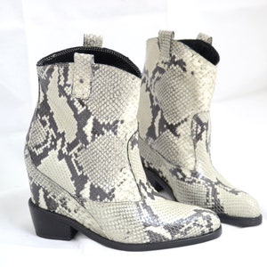 Puede incluir: Un par de botas vaqueras de piel de serpiente de color marrón claro y negro, con punta puntiaguda y tacón de cuña.