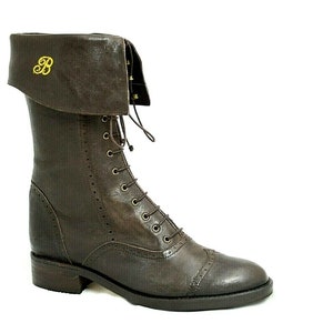Puede incluir: Botas de cuero marrón con un puño abatible y una letra "B" bordada en oro en el puño. Las botas tienen un cierre de cordones en la parte delantera y una puntera redondeada.