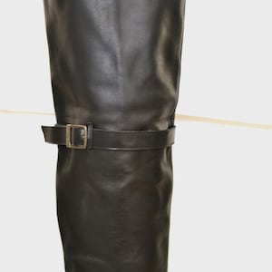 May include: Black leather knee-high boot with a buckle detail. The boot has a smooth texture and a simple design. The buckle is a gold color. The boot is a classic style.