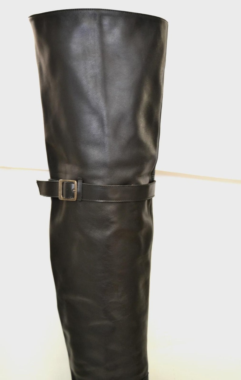 May include: Black leather knee-high boot with a buckle detail. The boot has a smooth texture and a simple design. The buckle is a gold color. The boot is a classic style.