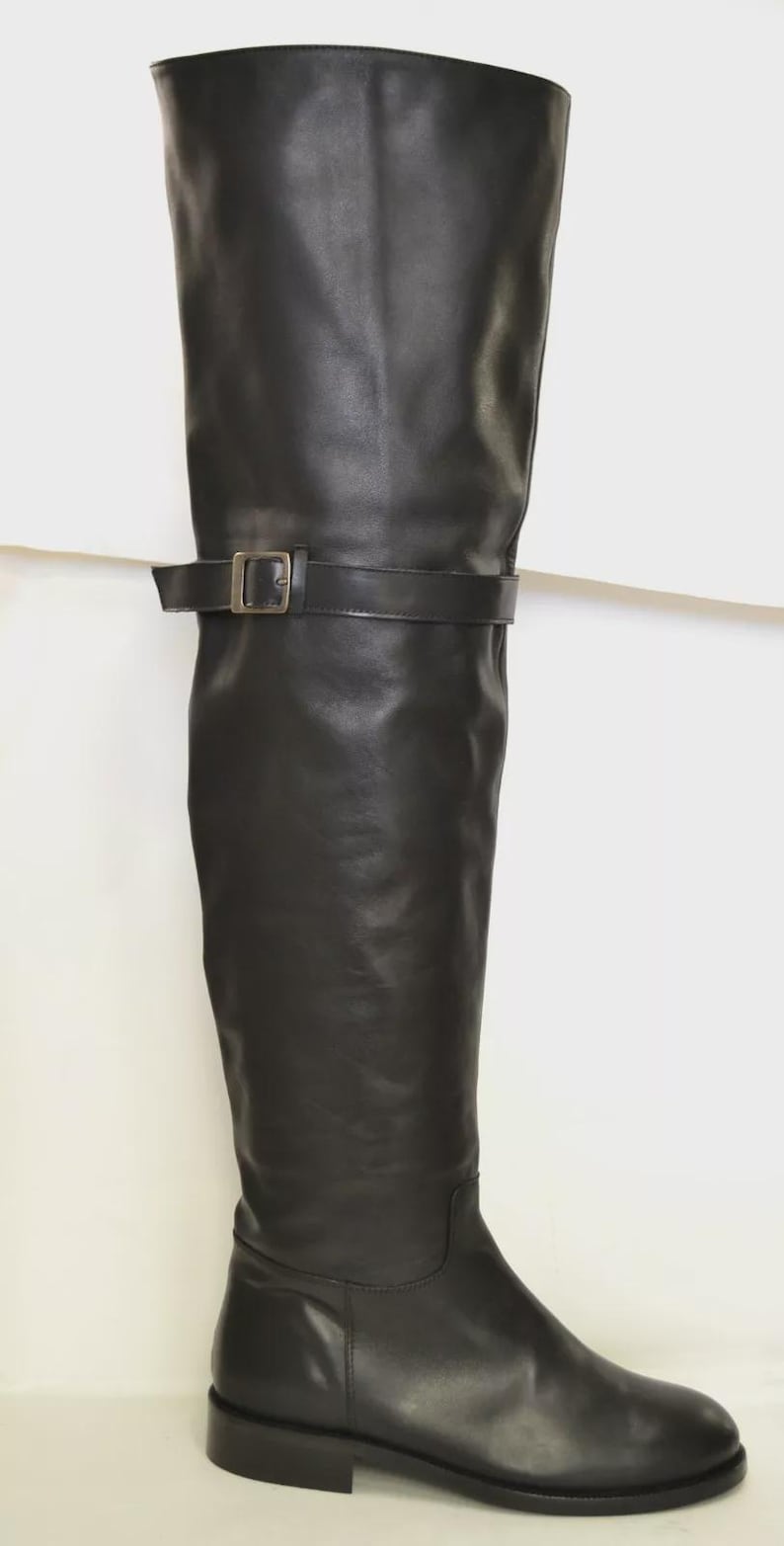 May include: A black leather knee-high boot with a buckle detail. The boot has a flat sole and a smooth, polished finish. The boot's shaft extends above the knee, and a strap with a silver buckle secures the boot.