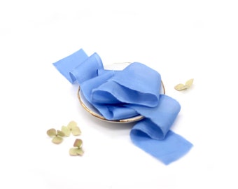 May include: A light blue silk ribbon on a white plate with a gold rim. The ribbon is arranged in a loose, flowing shape. There are a few small green leaves scattered around the plate.