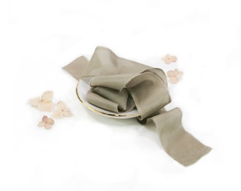 May include: A roll of sage green silk ribbon is draped over a white plate with a gold rim. The ribbon is textured and has frayed edges. Dried, light pink flowers are scattered around the plate, creating a soft, elegant aesthetic.