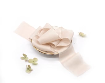 May include: A pale peach-colored silk ribbon is coiled on a small, gold-rimmed dish. The ribbon has a frayed edge and is partially unraveled. Scattered around the dish are small, dried, green flower buds, all set against a white background.