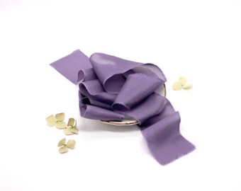 May include: A purple silk ribbon is displayed on a white plate with green flower petals scattered around it.