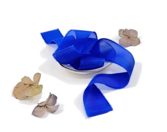 May include: A vibrant blue silk ribbon, artfully arranged on a small, silver-rimmed dish. The ribbon's texture is soft and slightly frayed, with a rich, saturated color. Dried flower petals in shades of beige and brown add a touch of natural contrast.