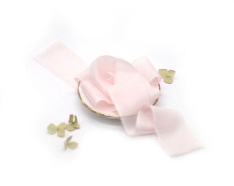 May include: A pale pink silk ribbon is arranged on a white surface with a small white plate and green flower petals.