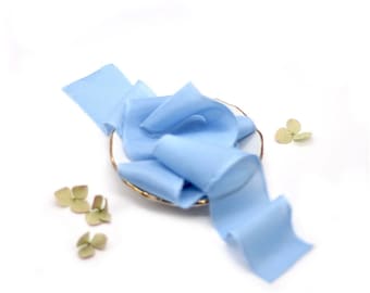 May include: A light blue silk ribbon is displayed on a small white plate with a gold rim. The ribbon is folded and twisted, creating a decorative display. There are small green flower buds scattered around the plate.