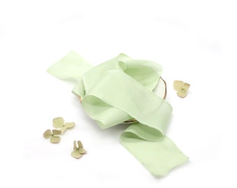 May include: A pale green silk ribbon is arranged on a white surface with small green flower buds scattered around it.