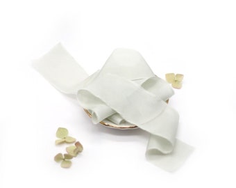 May include: A white silk ribbon is arranged in a decorative swirl on a small, gold-rimmed plate. The ribbon is soft and flowing, with a subtle sheen. There are a few dried green flower petals scattered around the plate.