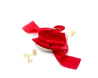 May include: A bright red silk ribbon is arranged on a white saucer. The ribbon is folded and twisted, creating a decorative display.