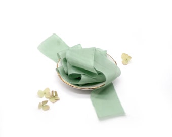 May include: A pale green silk ribbon is displayed on a small white plate with a gold rim. The ribbon is arranged in a loose coil and is surrounded by small green leaves.
