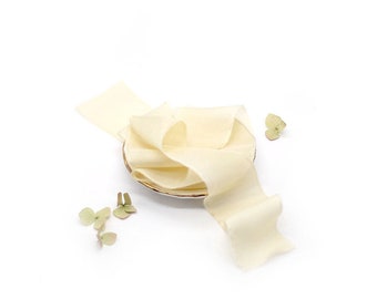 May include: A cream-colored satin ribbon is arranged on a small, gold-rimmed plate. The ribbon is folded and twisted, creating a decorative display. Small green leaves are scattered around the plate.