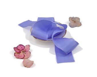 May include: A length of periwinkle blue silk ribbon with frayed edges, coiled on a small white dish with a gold rim. Dried flowers in shades of pink and beige are scattered around the ribbon, creating a delicate, artistic arrangement.