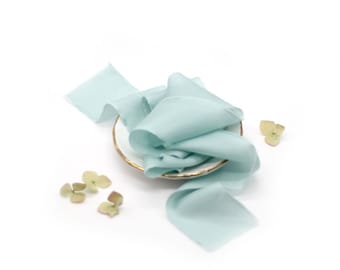 May include: A pale blue silk ribbon is arranged on a white plate with a gold rim. The ribbon is gathered in the center of the plate and flows out to the sides. There are small green leaves scattered around the plate.